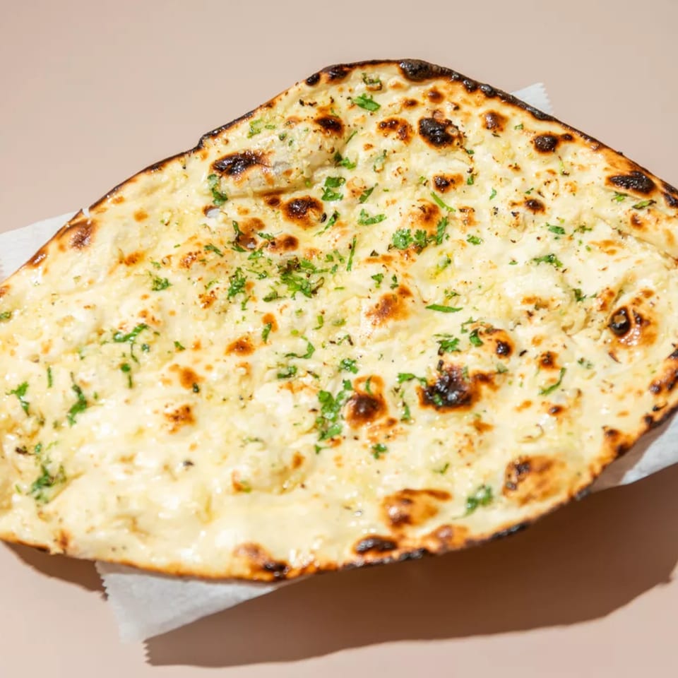 Close-up of a golden-brown garlic naan bread, topped with melted cheese and fresh green herbs.
