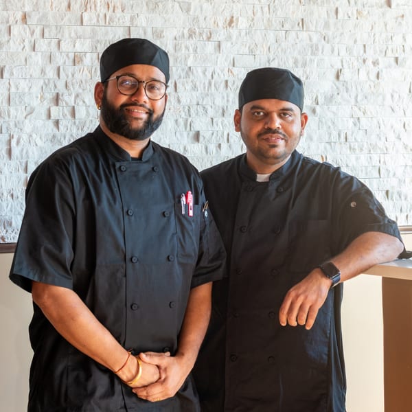 Two smiling Haldi Fine Indian Cuisine chefs in black uniforms stand against a white brick wall.